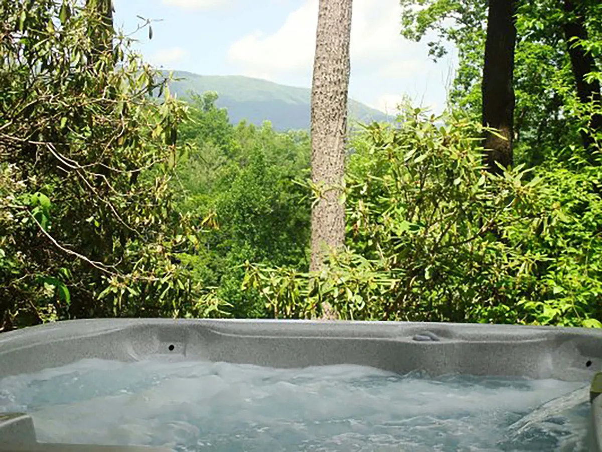 An inviting outdoor hot tub surrounded by lush greenery with a scenic mountain view in the background. The image conveys a peaceful atmosphere, ideal for relaxing in nature with friends after a day of golf or games.