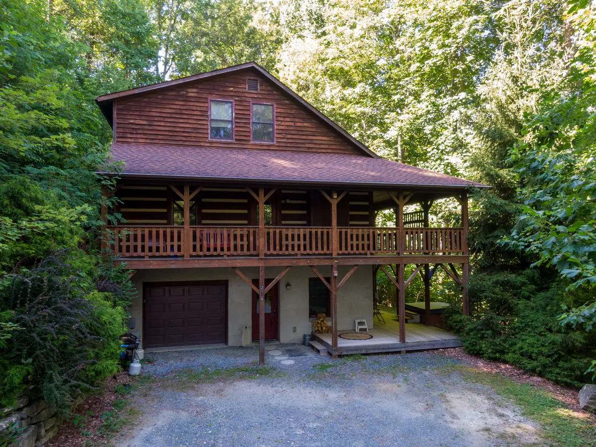 The hero image shows the inviting exterior of a three-story log cabin nestled in lush greenery, with a spacious wooden balcony overlooking the peaceful landscape. A stone pathway leads up to the entrance, and guests can imagine unwinding on the balcony at sunset or gathering with friends in the outdoor seating area. The scene exudes serenity and the promise of memorable moments in a mountain retreat.