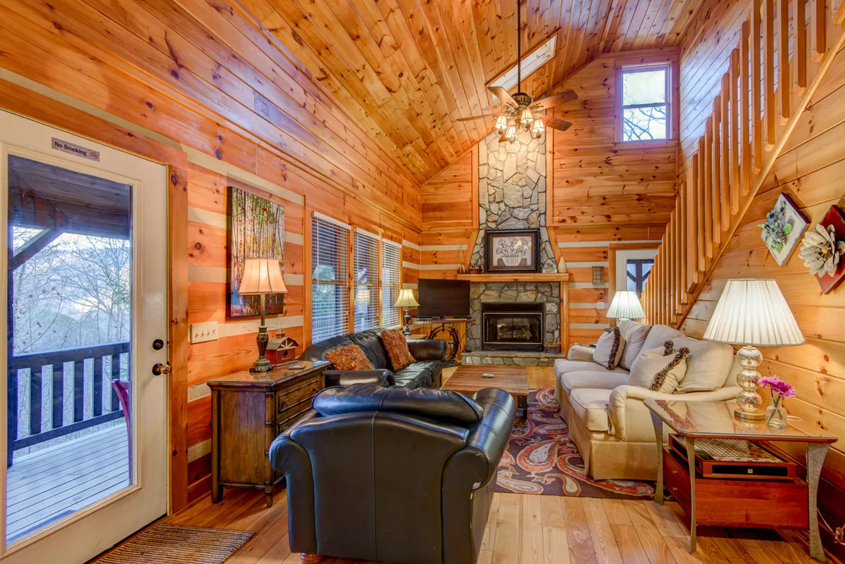 A welcoming lower-level den featuring a rustic stone fireplace, large windows letting in gentle light, a plush leather sofa, and soft armchairs. The ambiance is cozy and perfect for relaxing, watching TV, or enjoying warm fireside chats.