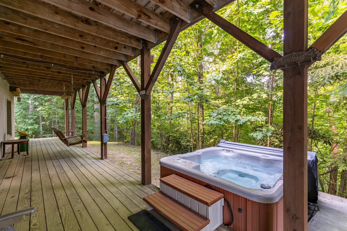 An exterior view of a wooden deck set amidst lush greenery, featuring a bubbling six-person hot tub. The deck offers space for socializing, soaking, and enjoying the tranquility of the surrounding woods.