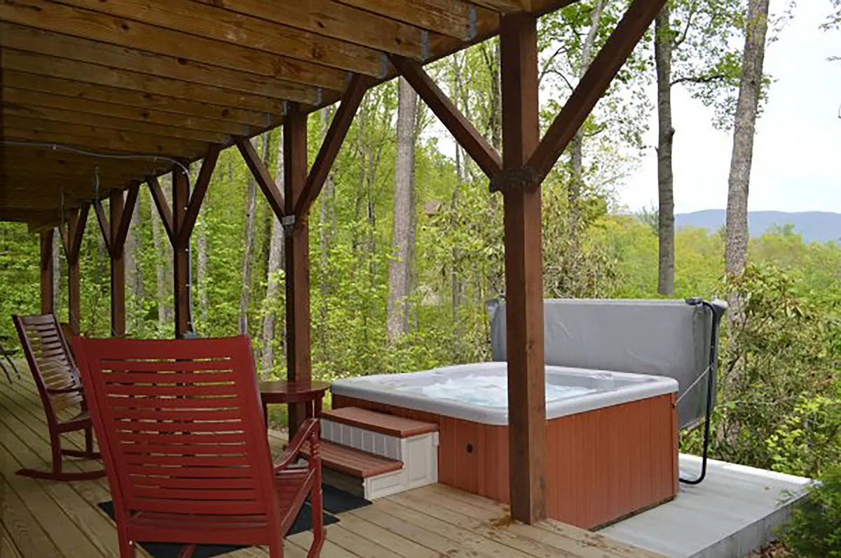 This image depicts an inviting exterior view of a cabin porch featuring rocking chairs and a hot tub, framed by sturdy wooden beams. The surrounding woods are visible, creating a sense of privacy and immersion in nature. The setting evokes relaxation and a connection to the tranquil, wooded landscape.