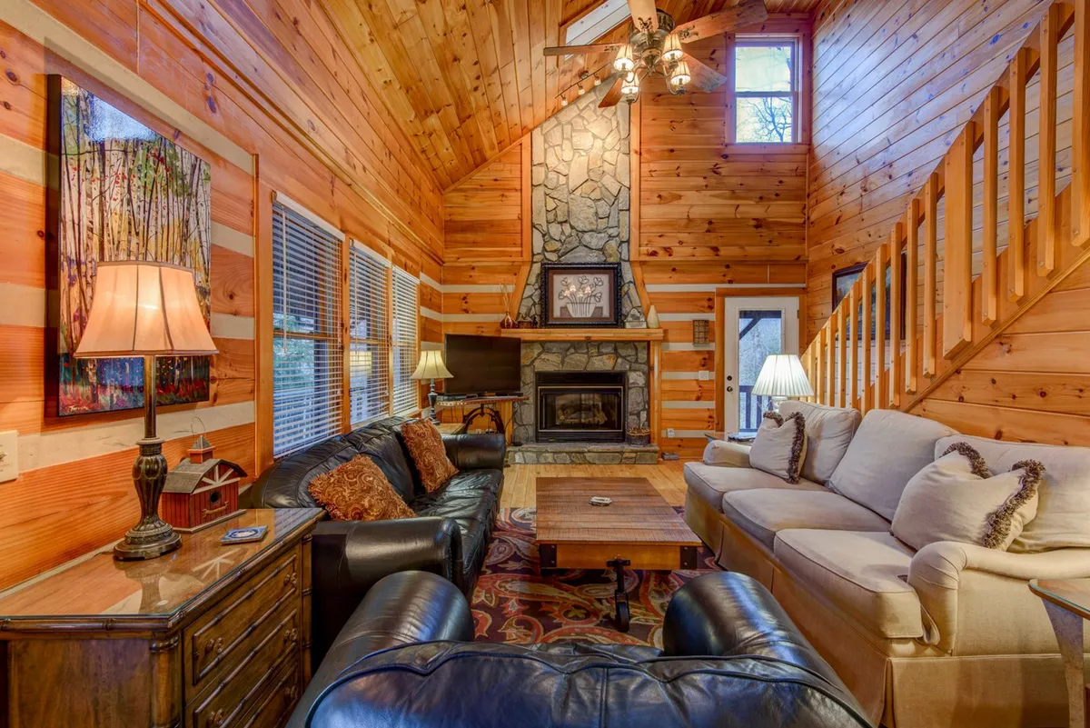 This image displays a spacious living room featuring a prominent stone fireplace, large windows that draw in natural light, sturdy wooden beams overhead, and a plush, inviting seating arrangement with comfortable sofas and armchairs. The atmosphere is warm and welcoming, perfect for gatherings by the fire or relaxed evenings with family and friends.