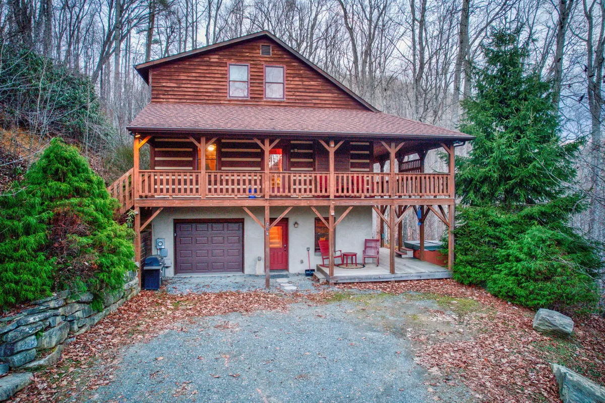 A serene exterior view of the cabin’s wooden balcony, front door, and rocky landscaping, surrounded by tall trees. The setting looks inviting for gatherings, evening chats, or enjoying the natural environment.