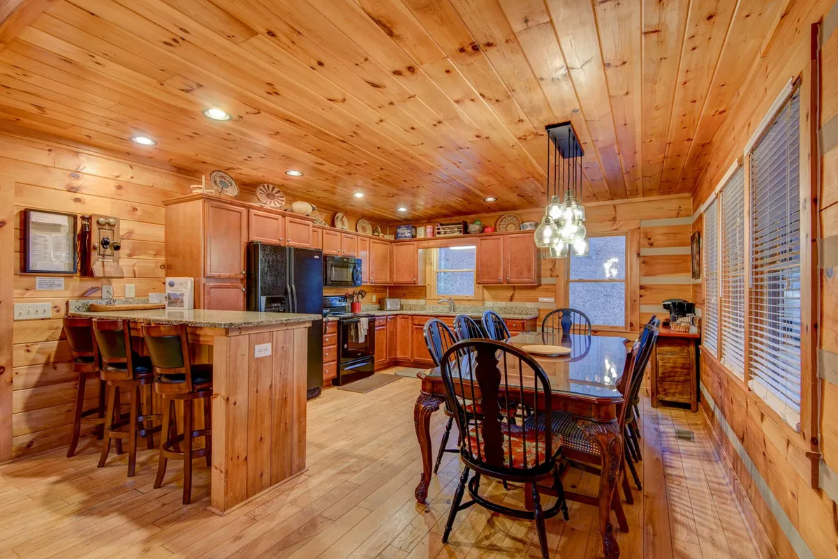 A bright, open-concept kitchen and dining area with a granite kitchen island, dining table with ample seating, wooden cabinetry, and large windows. The layout encourages communal cooking and shared meals, capturing the spirit of togetherness.
