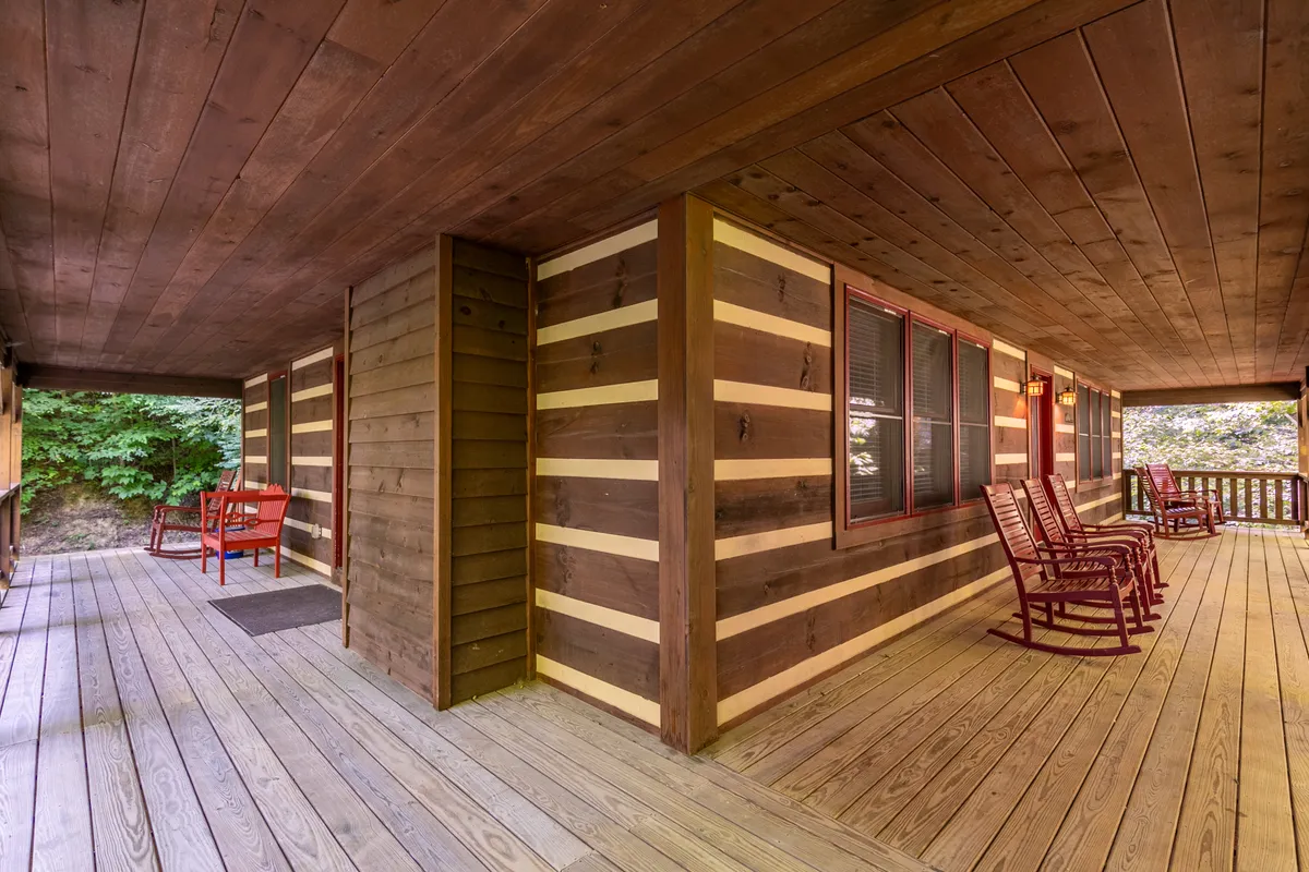 A cozy covered deck overlooking the woods, furnished with classic wooden rocking chairs and a red bench, set against the rustic log cabin walls. The deck looks out onto tranquil natural surroundings, offering a peaceful spot for morning coffee or evening relaxation.
