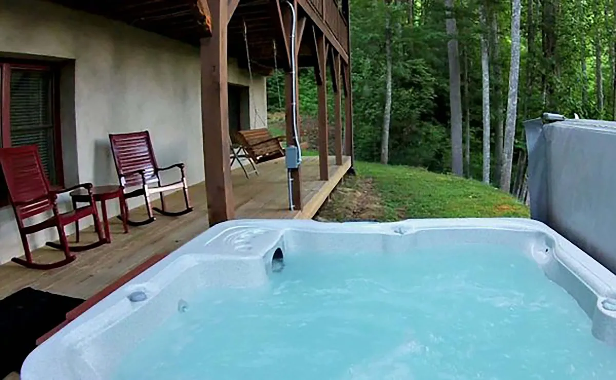 A tranquil exterior space featuring a hot tub and rocking chairs on the porch, surrounded by lush greenery and set up for relaxing while immersed in nature.